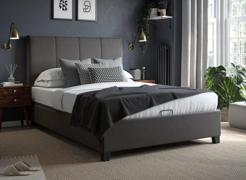 An upholstered grey ottoman bed frame with white bed sheets and grey accessories