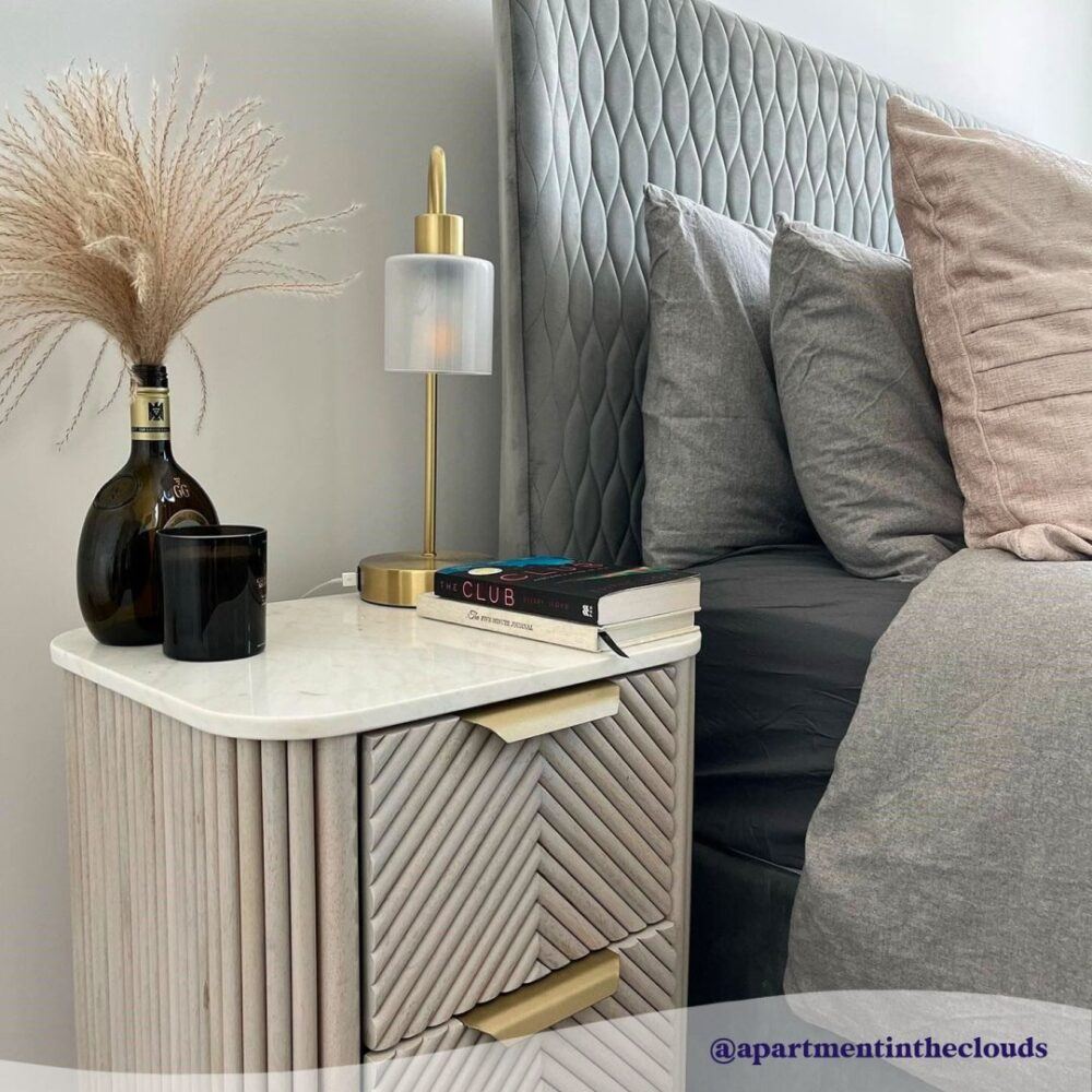 image of the side of a bed with a bedside table in shot on which are ornaments such as books, a table lamp.