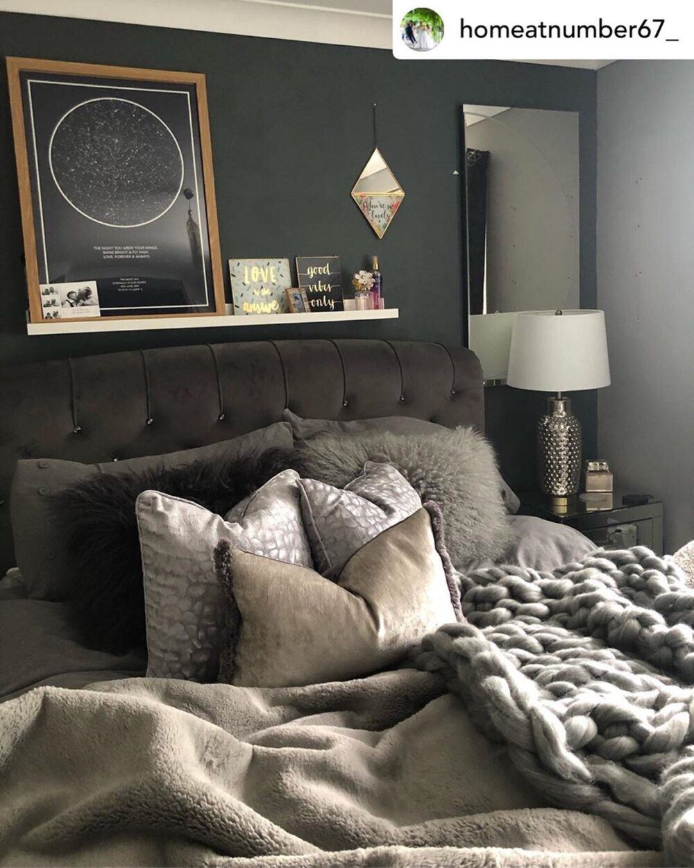 A dark and moody bedroom, using greys, greens and silver and gold touches to balance it out. A black headboard pairs with varying tones and textures of pillows, from fluffy to velvet, and a chunky knit throw matches a fleece one atop the bedsheets. Above it all, a floating shelf holds various cards and knickknacks.
