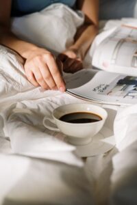 A cup of coffee is nestled between white sheets, while someone reads a book in bed.