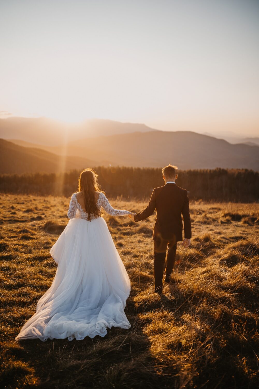 A couple walking over a field in wedding attire, with the sun rising in the distance.
