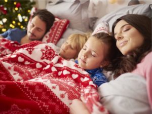 A family sleeping together in one bed with a red and white festive blanket and a christmas tree in the background