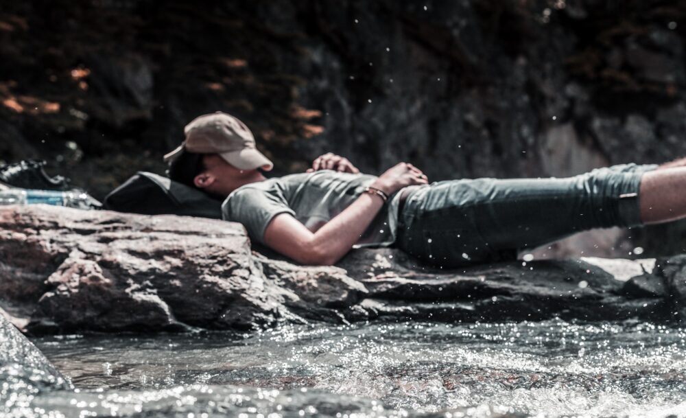 A man sleeps by water with a cap over his face