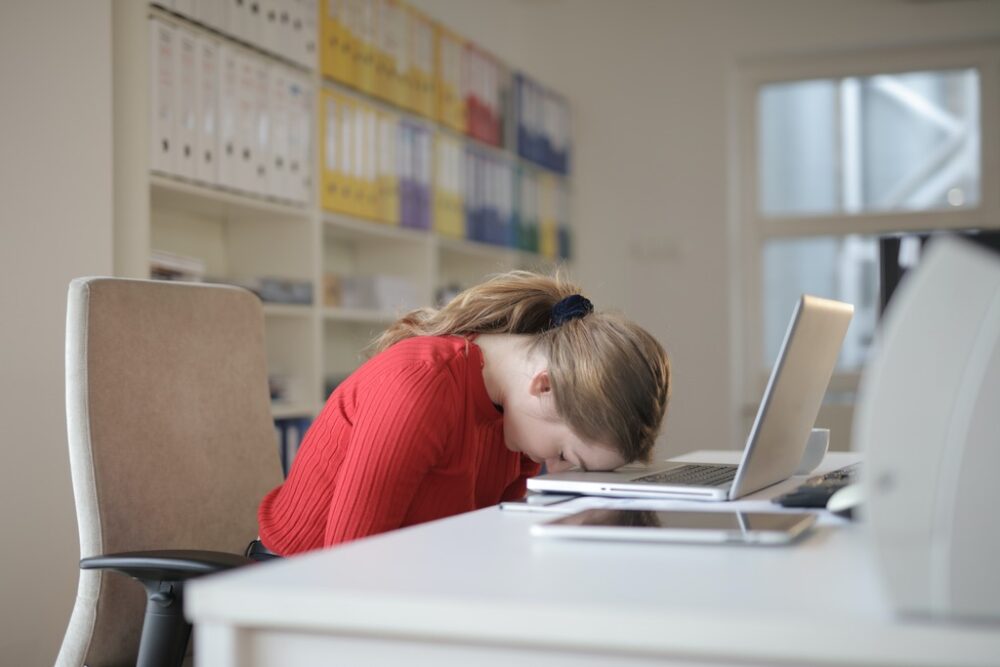A woman in a red shirt sleeping on her laptop at work.