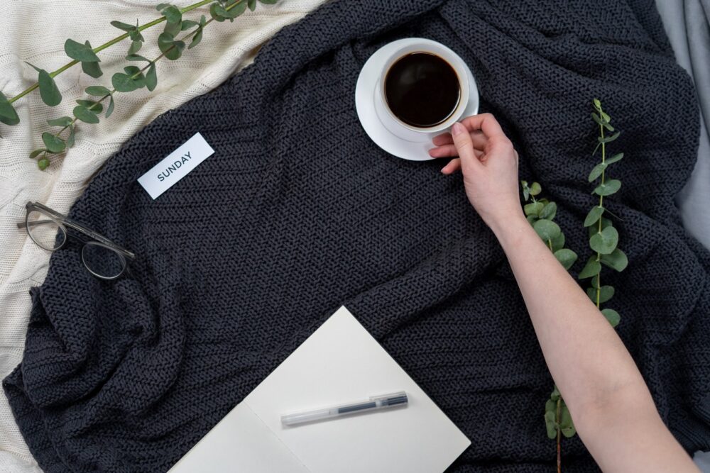 A hand reaches for a cup of coffee in a white cup on a jumper, surrounded with eucalyptus branches. A notecard says "sunday", and nearby is a pen and paper.