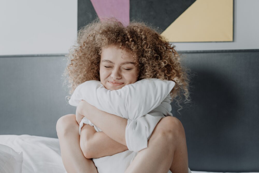 A young woman with curly hair sitting on bed hugging her white pillow, with a grey headboard and modern art behind her.
