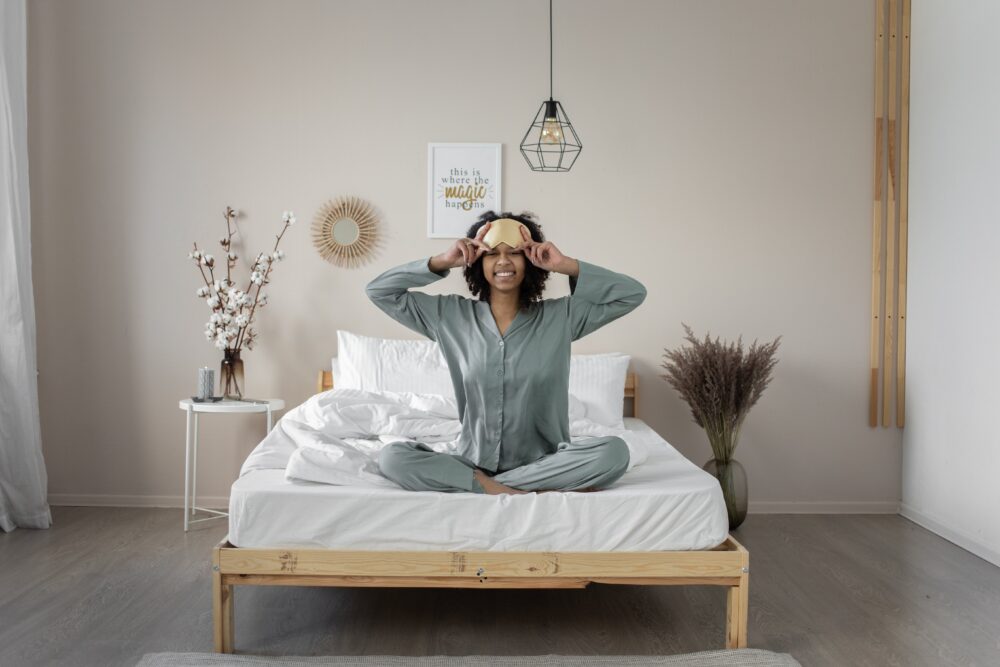 A woman wakes up in her bed looking happy and refreshed. She is wearing simple pyjamas, sitting up on the white sheeted bed with a wooden frame, and has her hands on a beige eyemask. Behind her is a minimalist decorated wall.
