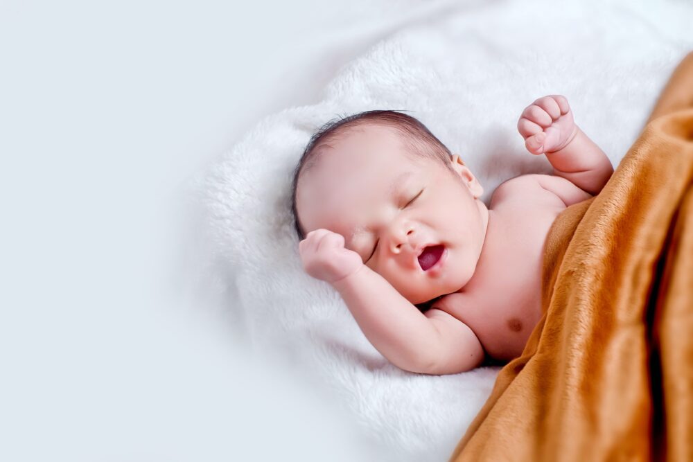 A baby waking up under a golden-yellow blanket, on a white pillow and white background.