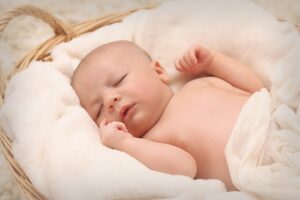 A baby sleeps in a wicker basket, surrounded by soft white blankets and cushions.