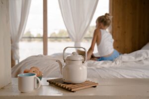 A white pot and mug sits on a wooden table, in front of a bed with a woman sitting on it.