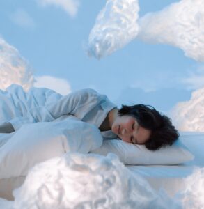 A person sleeps on a white sheet surrounded by fluffy clouds and a blue backdrop.