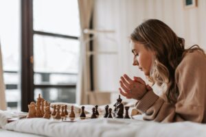 A woman playing chess in bed on white sheets with a bright white background.